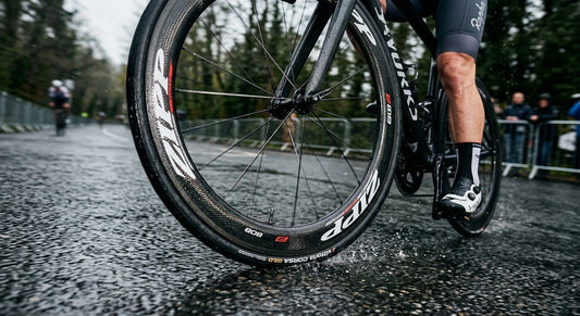 A close-up, high-speed action shot of a carbon fibre bike wheel rolling over a wet asphalt road, highlighting the sharp, seamless transition between the tyre and the rim sidewall.