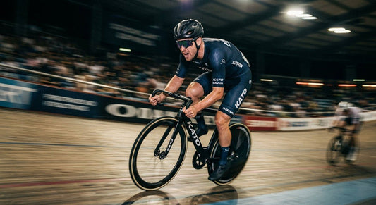 A professional cyclist wearing sleek Oakley QNTM Kato sunglasses during a high-speed sprint, captured with motion blur and dramatic stadium lighting.