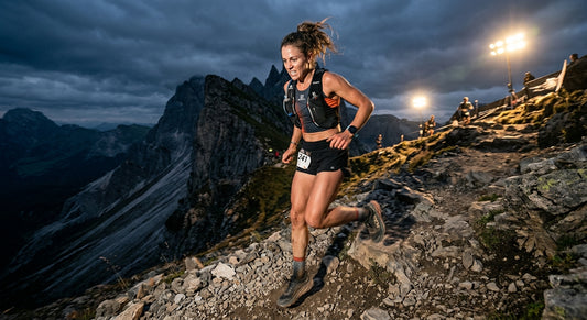 A professional trail runner mid-descent on a steep, rocky mountain path, capturing motion blur in the legs and intense concentration under dramatic stadium-style lighting.