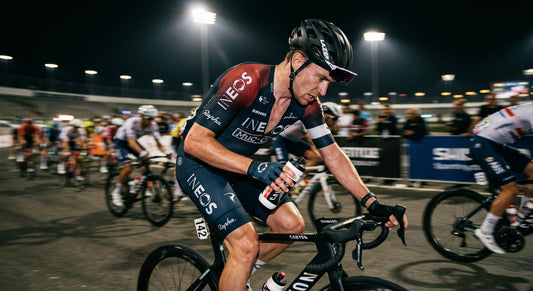 A close-up of a professional cyclist reaching for a bidon during a high-intensity road race with motion blur and dramatic stadium lighting.