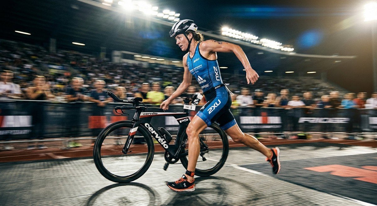 A professional triathlete in a blurred, high-speed motion shot transitioning from a racing bike to a running stride, captured with dramatic stadium lighting.