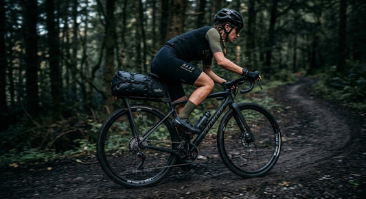 A sleek black Tailfin adventure rack mounted on a carbon gravel bike, set against a blurred forest trail background with moody, dramatic lighting.