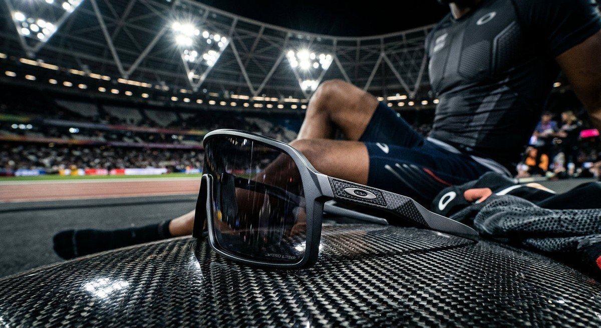 A sleek, futuristic pair of Oakley high-wrap sunglasses resting on a carbon fibre surface under dramatic stadium lighting.
