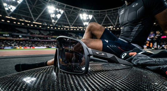 A sleek, futuristic pair of Oakley high-wrap sunglasses resting on a carbon fibre surface under dramatic stadium lighting.