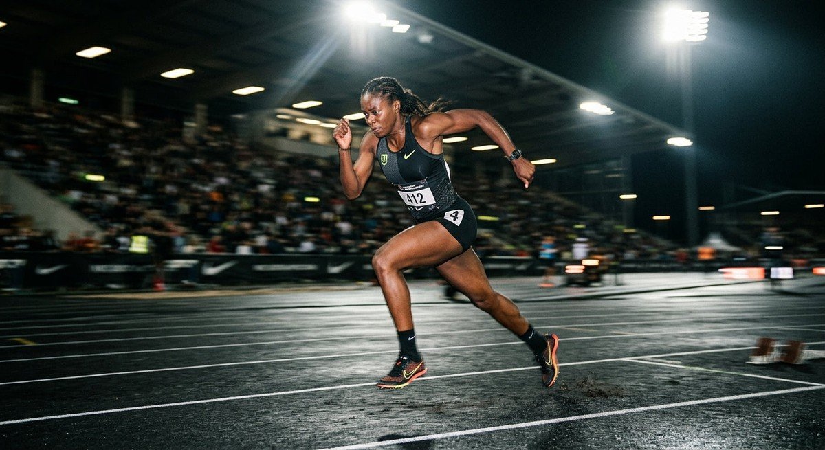 A professional athlete sprinting on a dark, moody track with dramatic stadium lighting and high-speed motion blur.