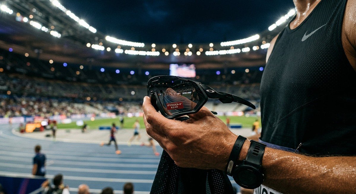 A close-up of high-performance sports sunglasses with sleek, aerodynamic lines held against a dramatic, moody stadium lighting background.