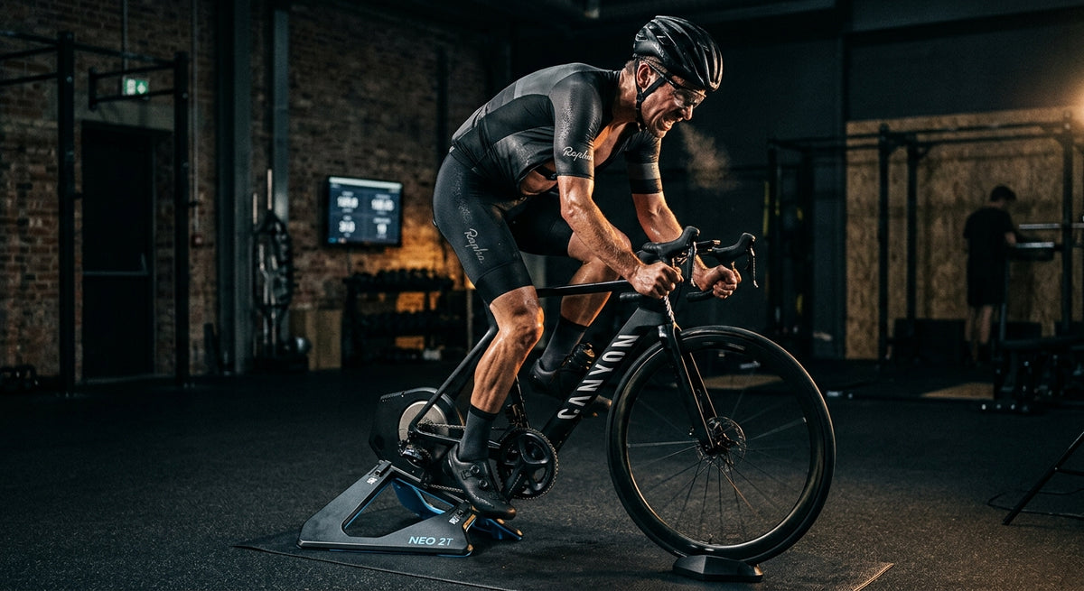 Professional cyclist pushing hard on an indoor smart trainer in a dark, moody gym with dramatic cinematic lighting.