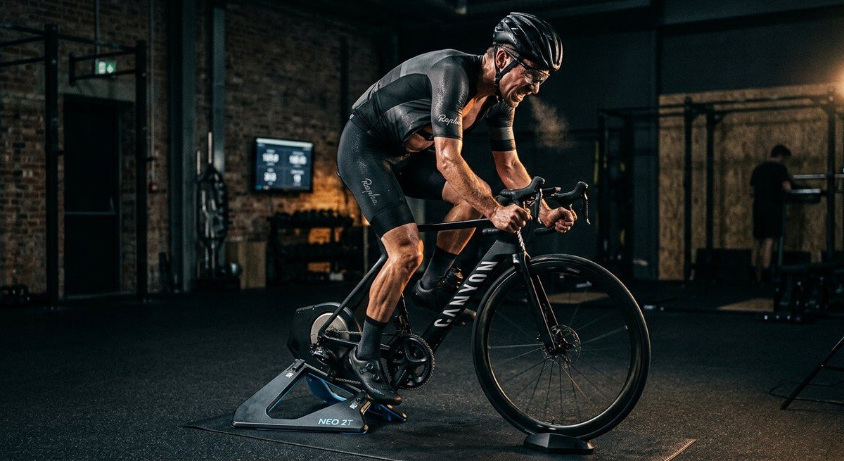 Professional cyclist pushing hard on an indoor smart trainer in a dark, moody gym with dramatic cinematic lighting.