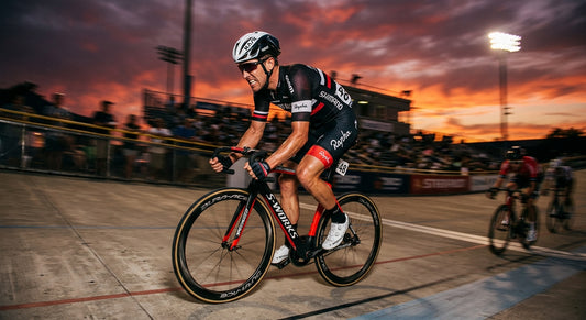 A professional road cyclist sprinting with intense focus, captured in high-speed motion blur against a dramatic, moody sunset background at a velodrome.