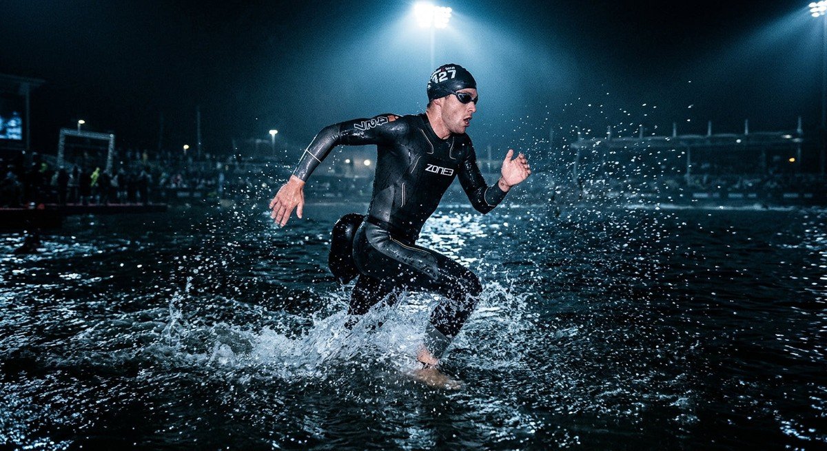 A professional triathlete in a high-performance neoprene wetsuit sprinting through dark, moody open water with cinematic stadium lighting illuminating the water droplets.