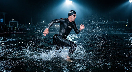 A professional triathlete in a high-performance neoprene wetsuit sprinting through dark, moody open water with cinematic stadium lighting illuminating the water droplets.