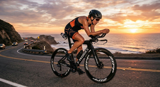 A professional triathlete in a sleek aerodynamic position on a high-end carbon fibre bike during a sunset training session.