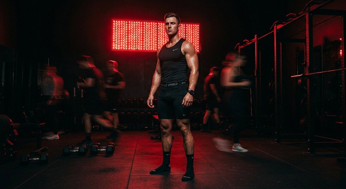 A professional athlete standing in front of a high-power red light therapy panel in a dark, moody gym with dramatic cinematic lighting and motion blur.