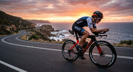 A triathlete in a low aero position cycling at high speed on a coastal road at dawn.