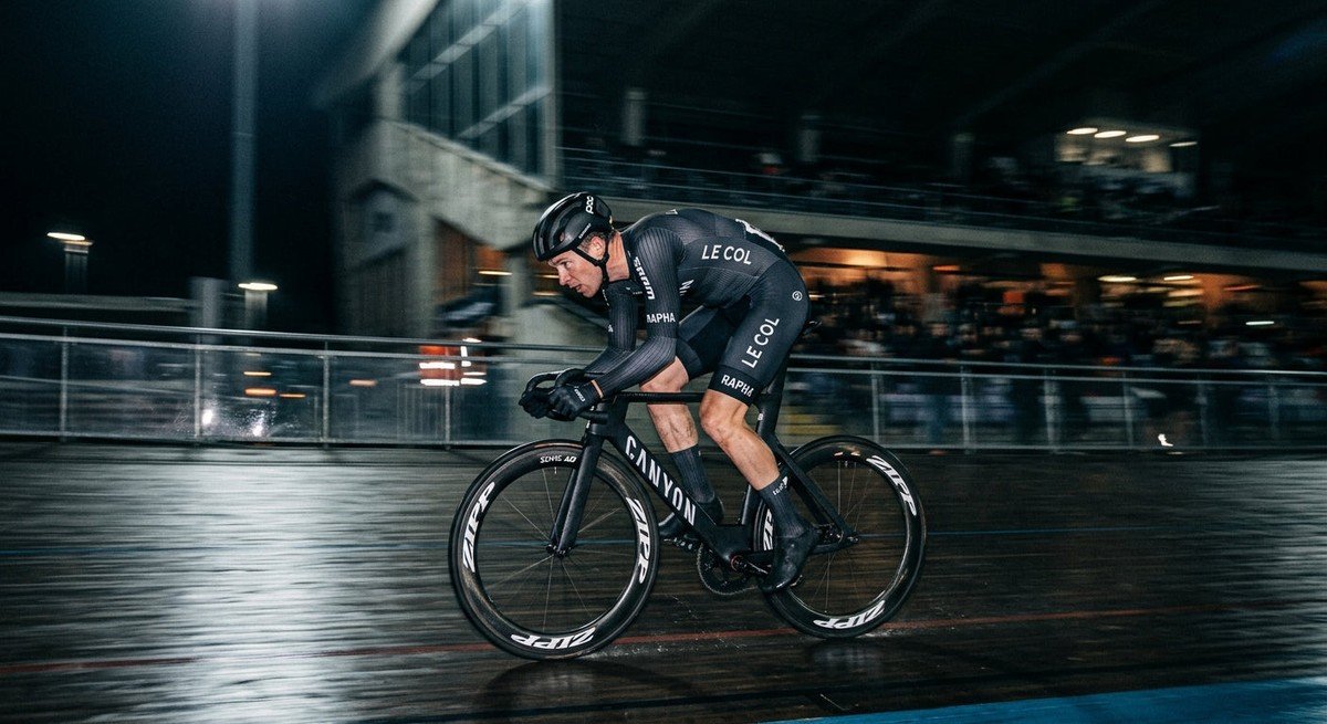 A professional cyclist in a tuck position on a carbon fibre bike, captured in high-speed motion blur against a moody, dark stadium background.