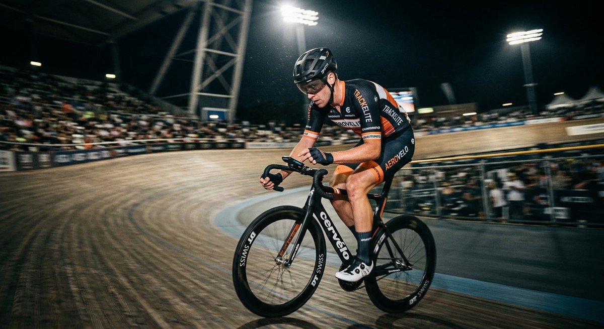 A professional cyclist in a blurred motion shot, illuminated by dramatic stadium lights, checking a digital device while riding on a velodrome track.