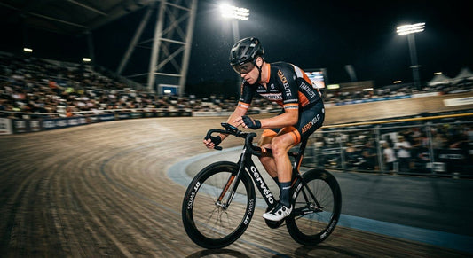 A professional cyclist in a blurred motion shot, illuminated by dramatic stadium lights, checking a digital device while riding on a velodrome track.