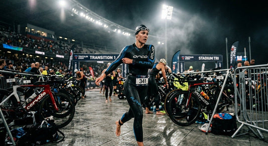 A professional triathlete in a sleek wetsuit rushing through a transition area in a stadium with dramatic, moody lighting.