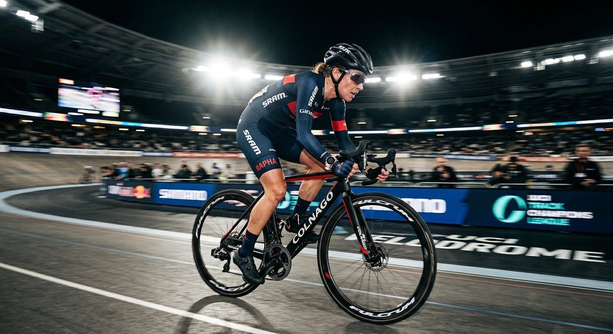 A professional cyclist in a focused athletic stance, backlit by dramatic stadium lighting with a sharp motion blur effect.