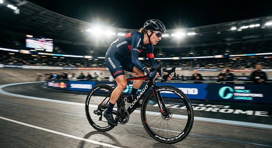 A professional cyclist in a focused athletic stance, backlit by dramatic stadium lighting with a sharp motion blur effect.