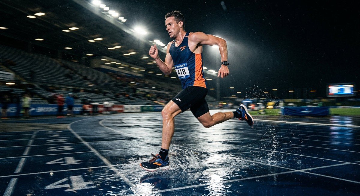 A professional action shot of a runner testing the Brooks Ghost 18 on a wet track with dramatic stadium lighting and high-speed motion blur.