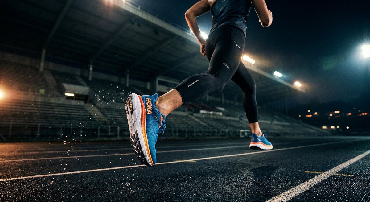 A sleek Hoka Skyward X2 running shoe captured in high-speed motion against a dark, moody stadium backdrop with cinematic lighting.