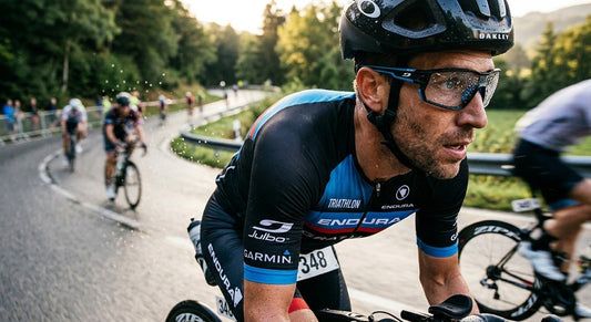 A close-up action shot of a triathlete wearing Julbo Edge sunglasses, with sharp water droplets on the lens and motion blur of the road in the background.