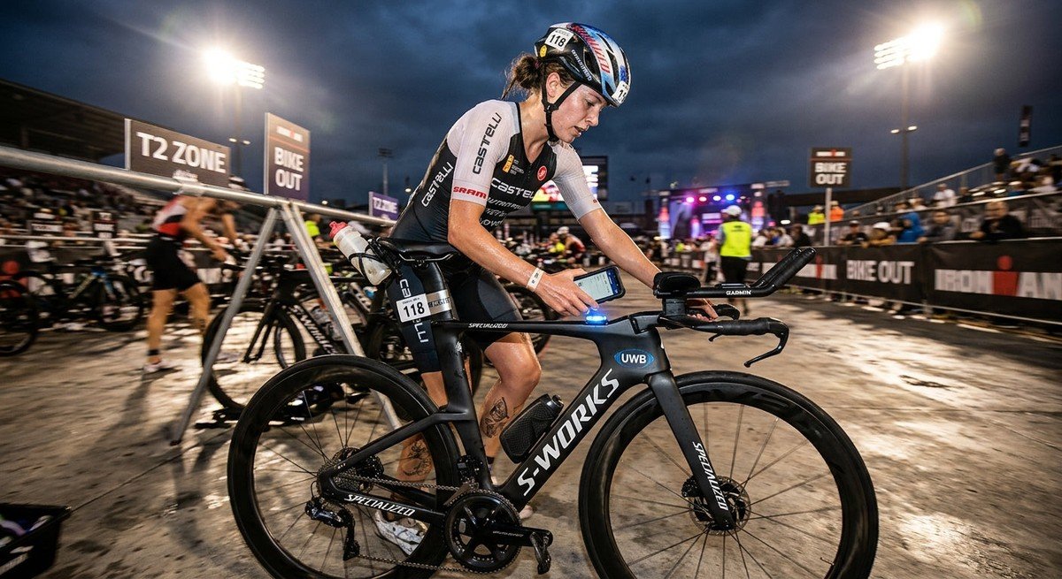 A professional triathlete in a high-speed transition zone, using a smartphone to scan a UWB-enabled tracking tag on a carbon fibre bike frame under dramatic cinematic stadium lighting.