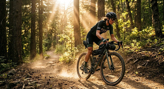 A professional cyclist in a sleek jersey riding a gravel bike through a dusty, sun-drenched forest path.