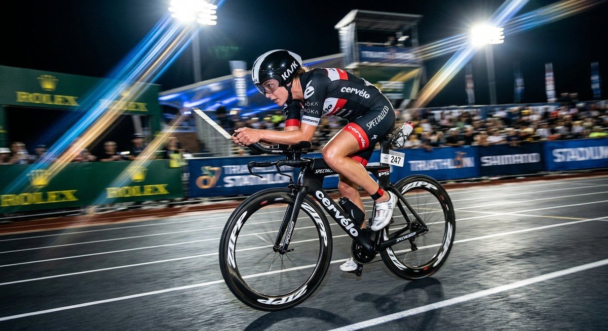 A professional triathlete in an aerodynamic tuck position on a carbon fibre bike, captured with motion blur and dramatic stadium lighting.