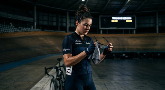 A professional athlete wiping down high-performance cycling sunglasses with a microfibre cloth in a moody, cinematic stadium environment.