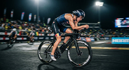 A professional triathlete in a blurred, high-speed cycling motion, demonstrating optimal aerodynamic form under moody, cinematic stadium lighting.