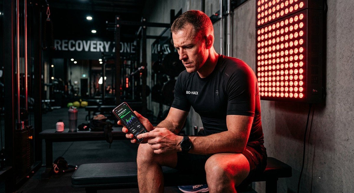 A biohacker analysing recovery data on a smartphone while illuminated by the intense, deep red glow of a professional photobiomodulation device in a modern athletic facility.