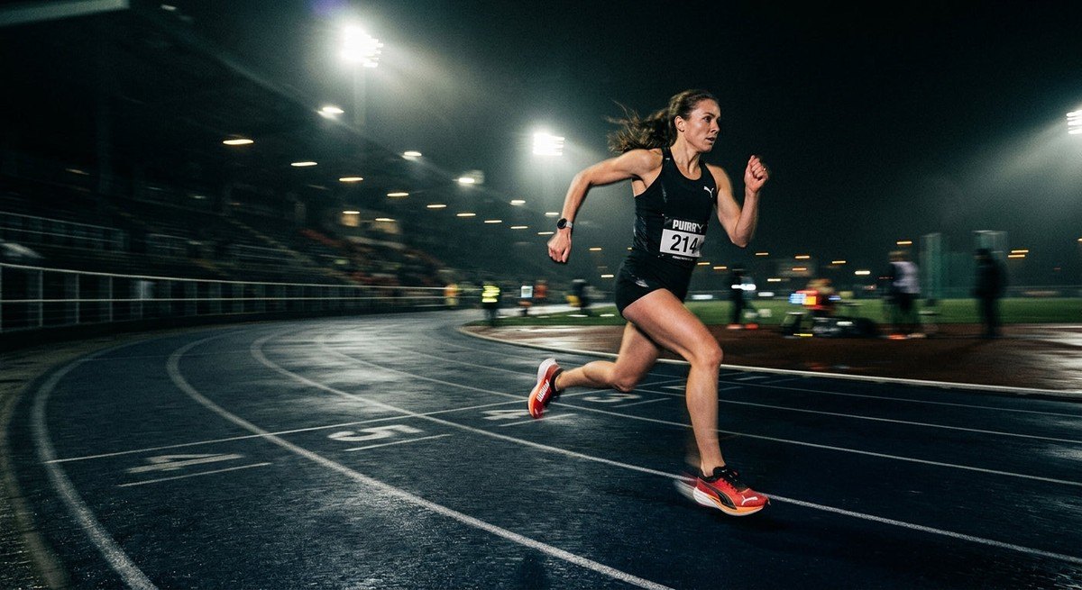 A blurry, motion-filled photograph of a runner wearing the Puma Deviate Nitro Elite 4 on a dark track under stadium floodlights.