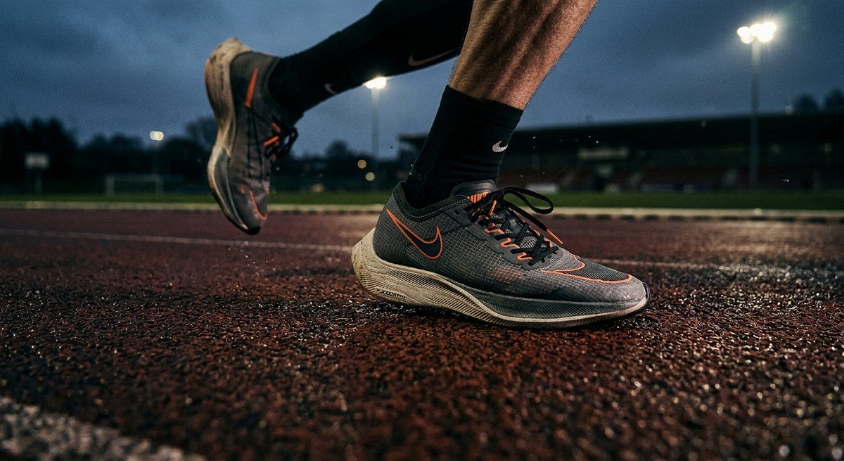 A close-up of a runner's feet hitting the track, showing sharp detail of the running shoe, texture of the track surface, and cinematic moody lighting.