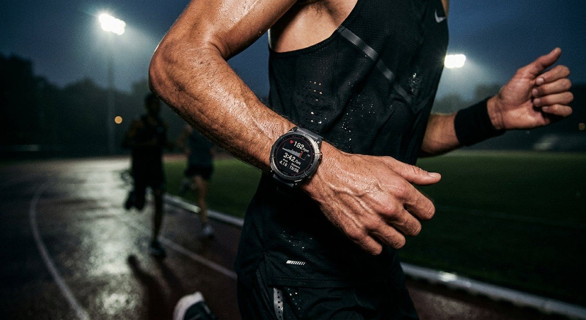 A close-up of a runner's smartwatch displaying heart rate and pace metrics during an intense track session under cinematic moody lighting.