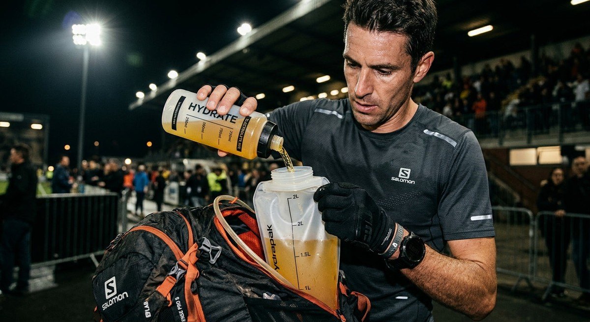 A close-up shot of an athlete filling a hydration bladder with a precise liquid mixture, dramatic stadium lighting highlighting the texture of the gear.