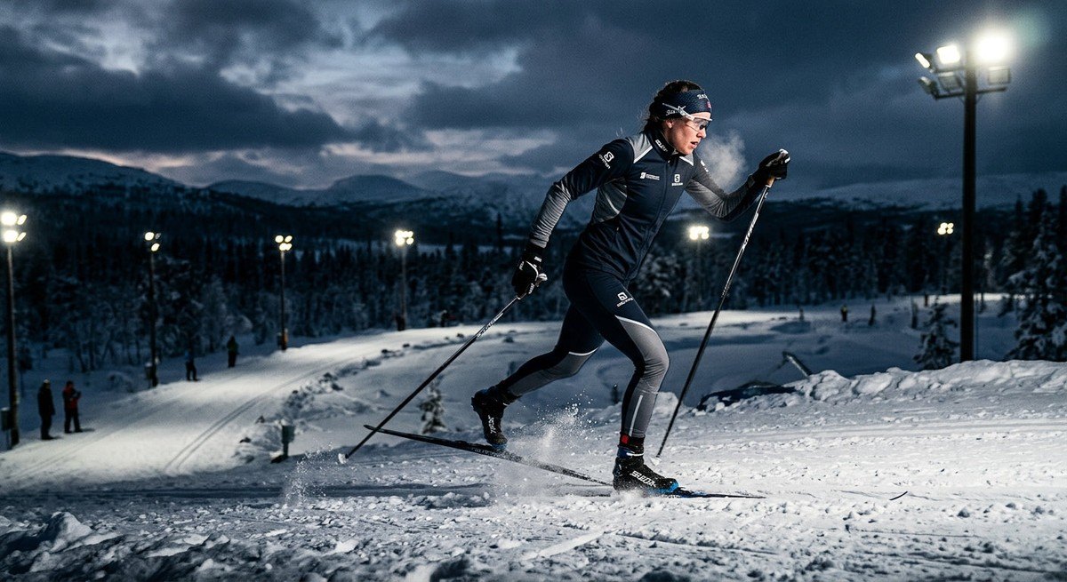 A cross-country skier in dynamic motion across a frozen landscape, captured with a moody, high-contrast aesthetic and stadium-style lighting.
