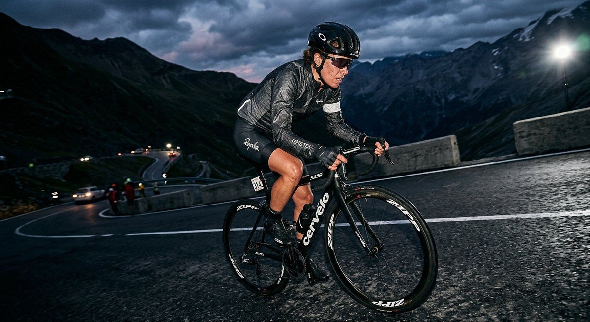 A cyclist in a high-tech windbreaker pushing hard during a steep mountain climb, captured with sharp focus and intense, moody stadium-style lighting.