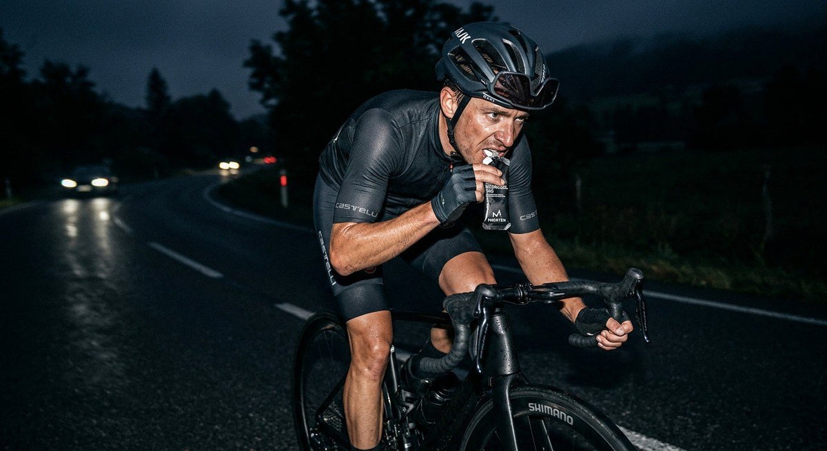 A cyclist taking a hydrogel during a high-intensity training session, with sharp focus on the athlete's concentrated expression and the aerodynamic gear, set against a dark, cinematic background.