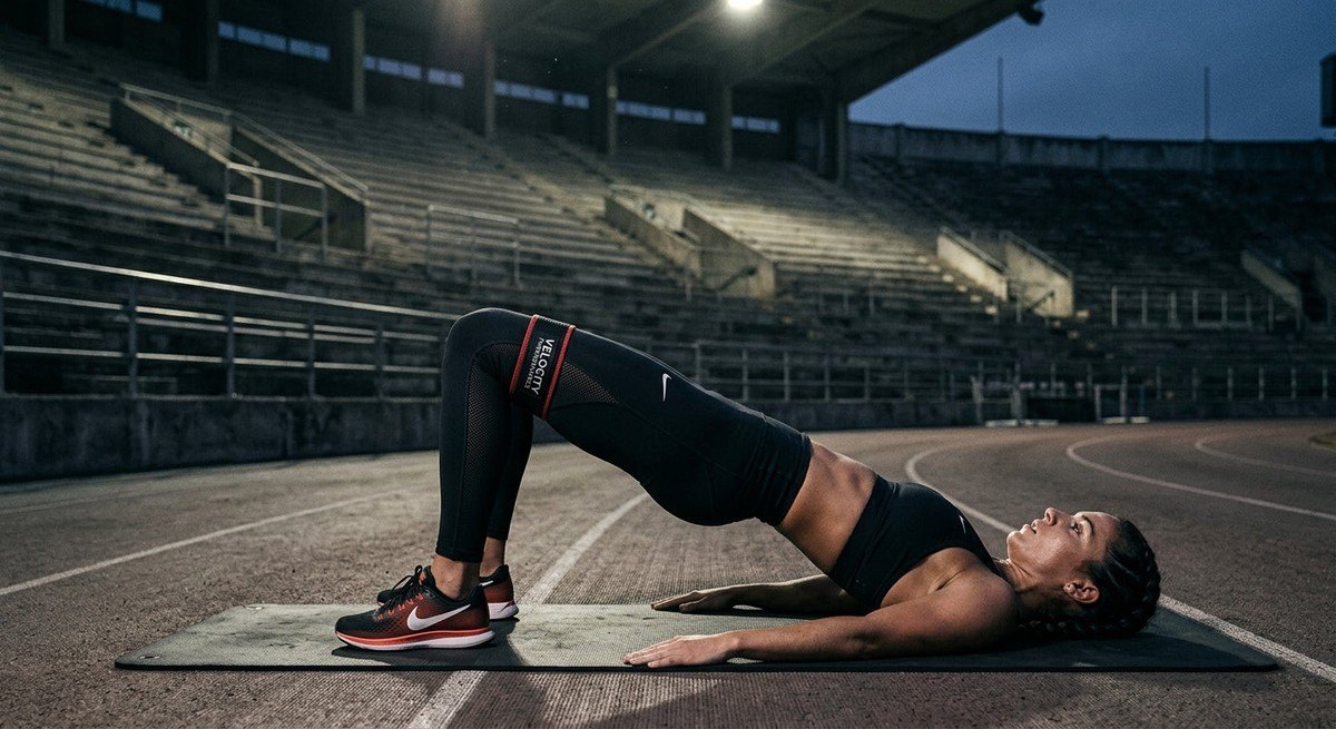 A high-speed action shot of a runner preparing for a workout, with a professional resistance band visible during a glute bridge exercise in a moody stadium setting.
