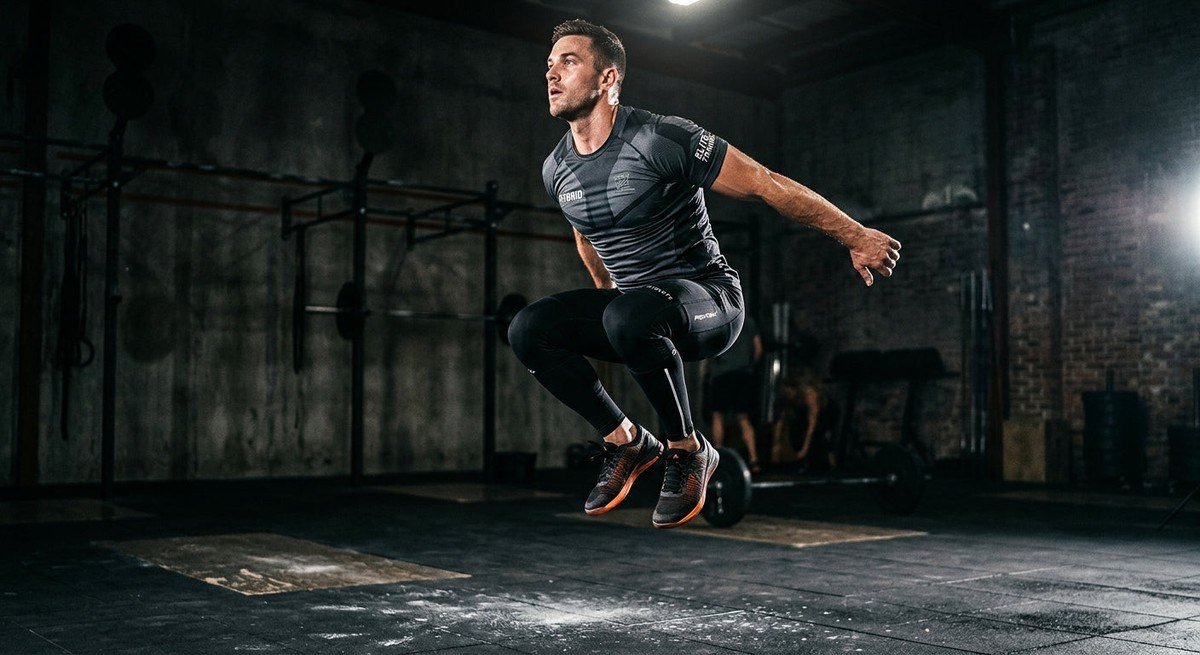 A hybrid athlete performing an explosive jump, wearing technical performance leggings and a tee, set against a dark, high-contrast background.