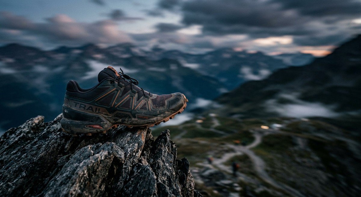 A moody, dramatic side profile of a high-performance trail shoe resting on a jagged mountain peak, with soft-focus motion blur in the background.