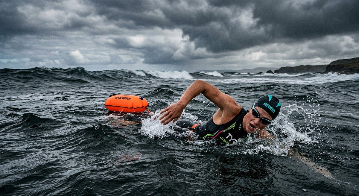 A professional swimmer navigating through choppy, dark open water with a bright orange tow float visible in the background under heavy, cinematic clouds.