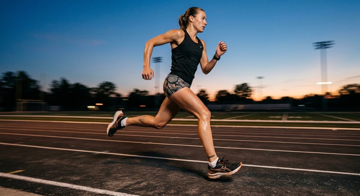 A side-profile shot of an athlete running on a paved outdoor track during twilight with motion blur and sharp focus on the foot strike.