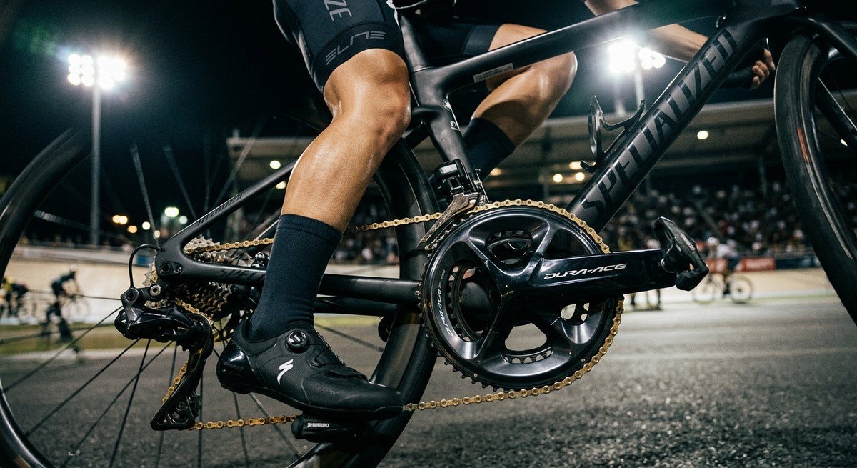 A sleek, carbon-fibre road bike crankset and rear derailleur illuminated by dramatic stadium lighting, emphasising metallic textures and professional quality.