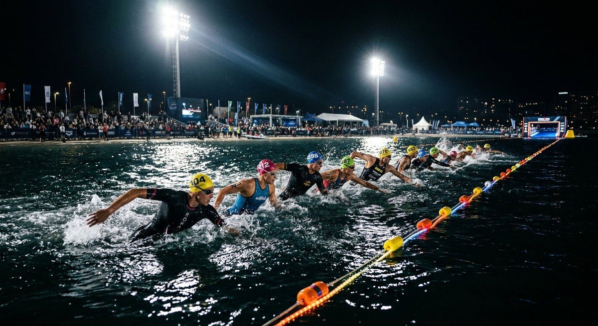 A wide-angle cinematic view of a group of triathletes swimming in a straight line during an open water race under intense stadium lighting.