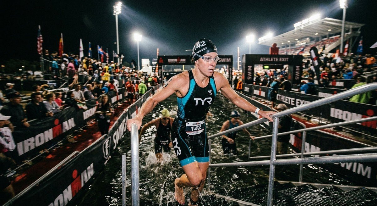 A wide-angle, moody shot of a swimmer emerging from the water at a stadium-lit triathlon event, motion blur capturing the intensity of the race.
