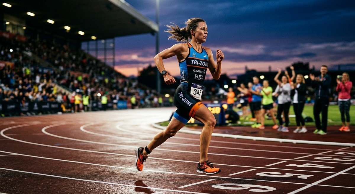 Action photography of a triathlete running on a track at dusk, highlighting the athletic form and realistic motion blur in a stadium setting.