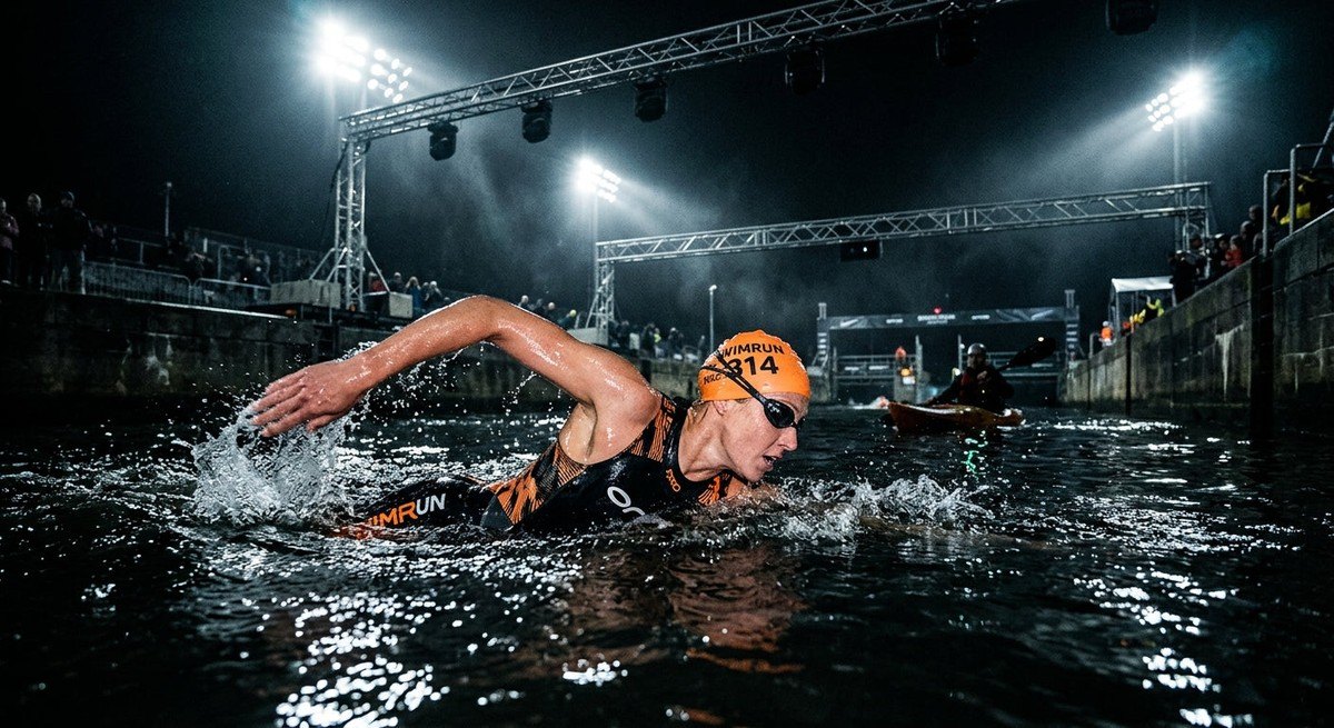 An athlete in specialised swim-run gear mid-stroke in a dramatic, dark-water channel with stadium-style lighting overhead.